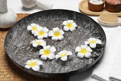 Photo of Bowl of water with plumeria flowers, herbal bags, towel and brush on table. Spa treatment