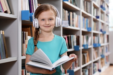Cute little girl with headphones reading book in library