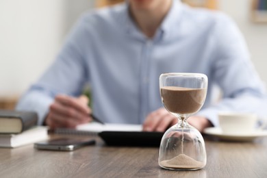 Photo of Hourglass with flowing sand on desk. Man taking notes while using calculator indoors, selective focus