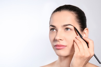 Photo of Young woman correcting shape of eyebrow with brush on light background