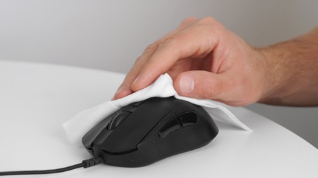 Man cleaning computer mouse with disinfecting wipe at white table indoors, closeup. Protective measures