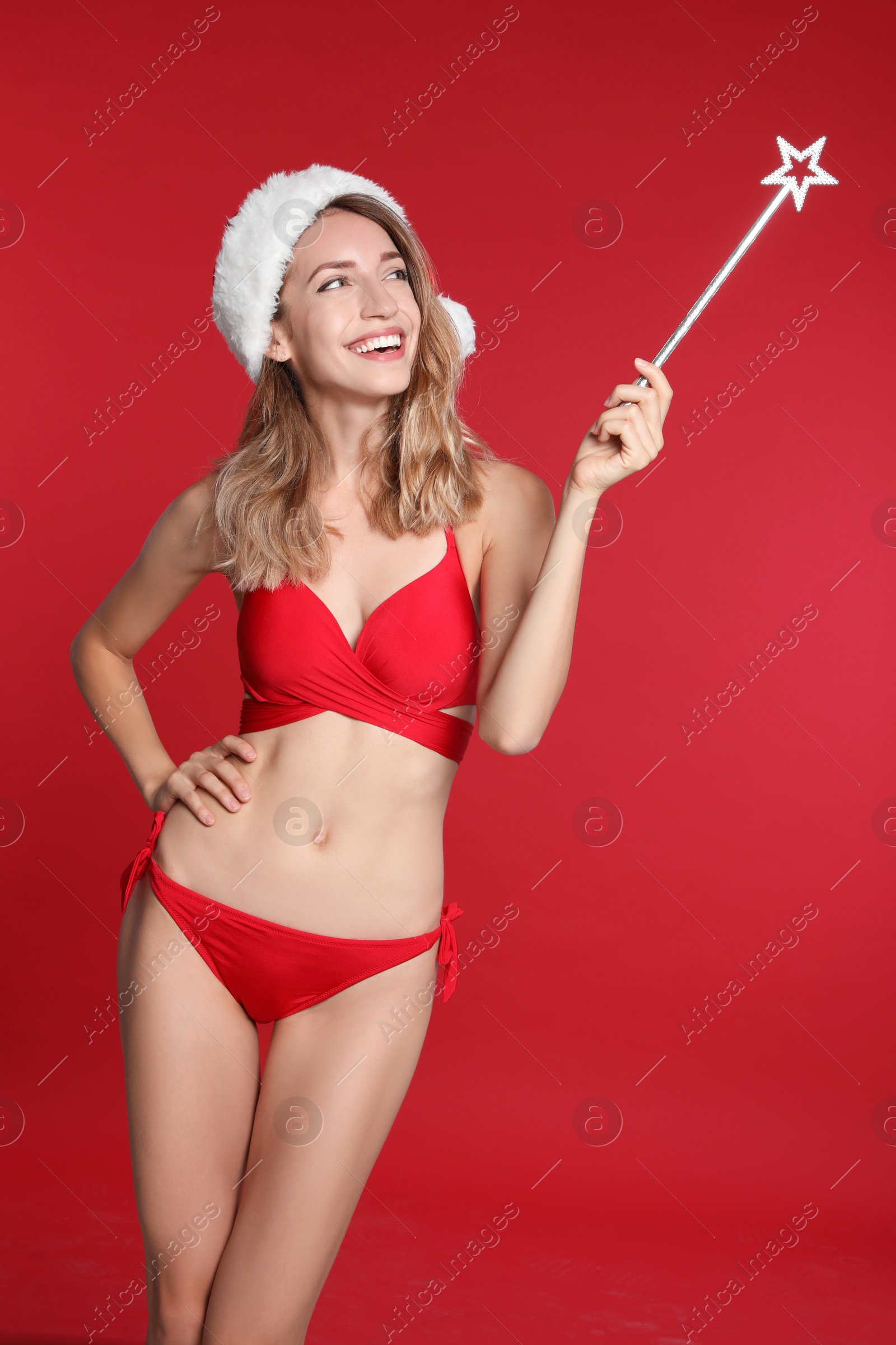 Photo of Sexy young woman in bikini and Santa hat with magic stick on red background. Christmas celebration