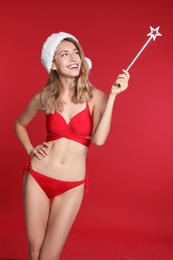 Photo of Sexy young woman in bikini and Santa hat with magic stick on red background. Christmas celebration