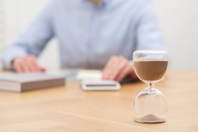 Photo of Hourglass with flowing sand on desk. Man using calculator indoors, selective focus