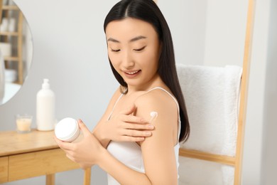 Photo of Beautiful young Asian woman applying body cream on shoulder in room