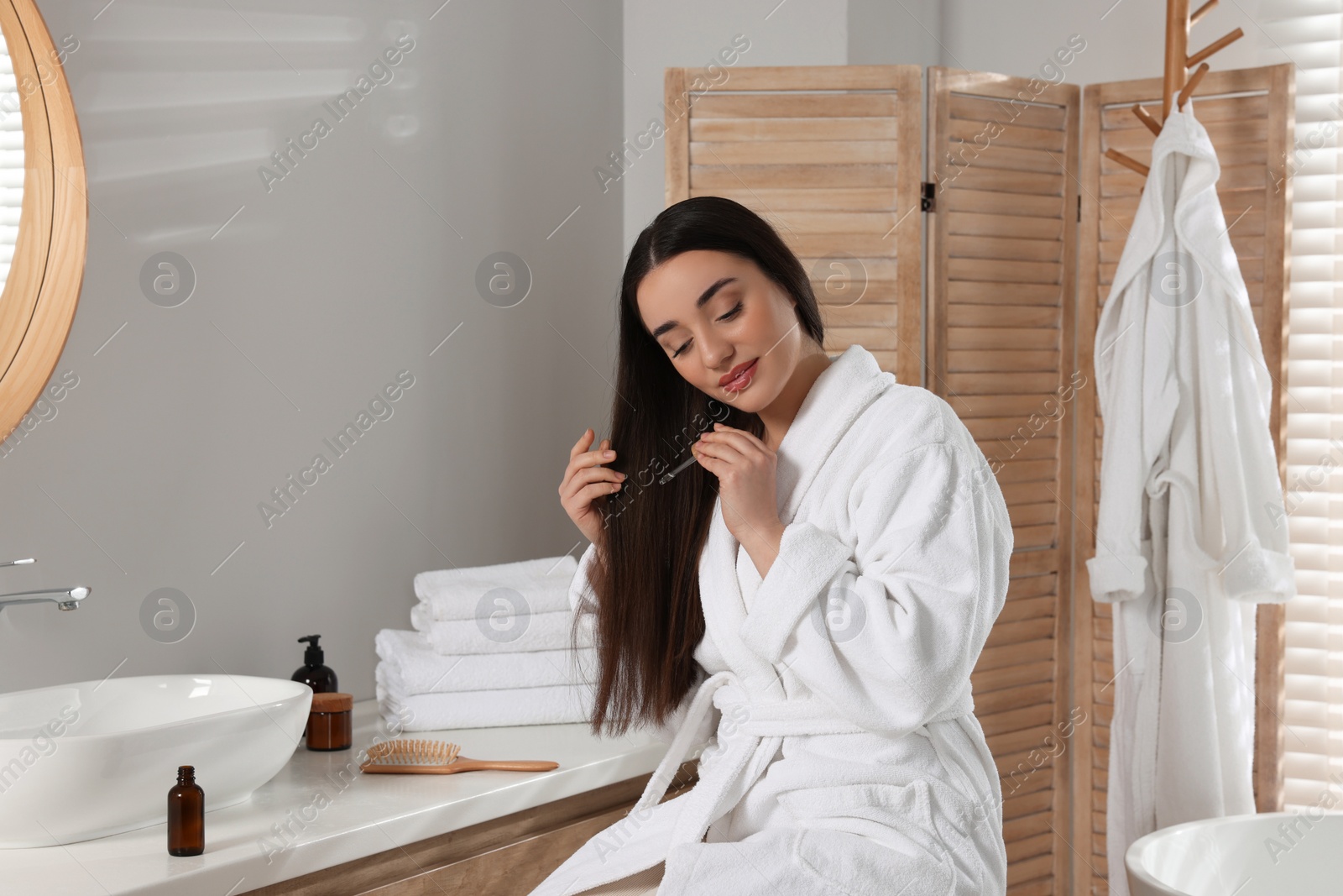 Photo of Young woman applying essential oil onto hair in bathroom