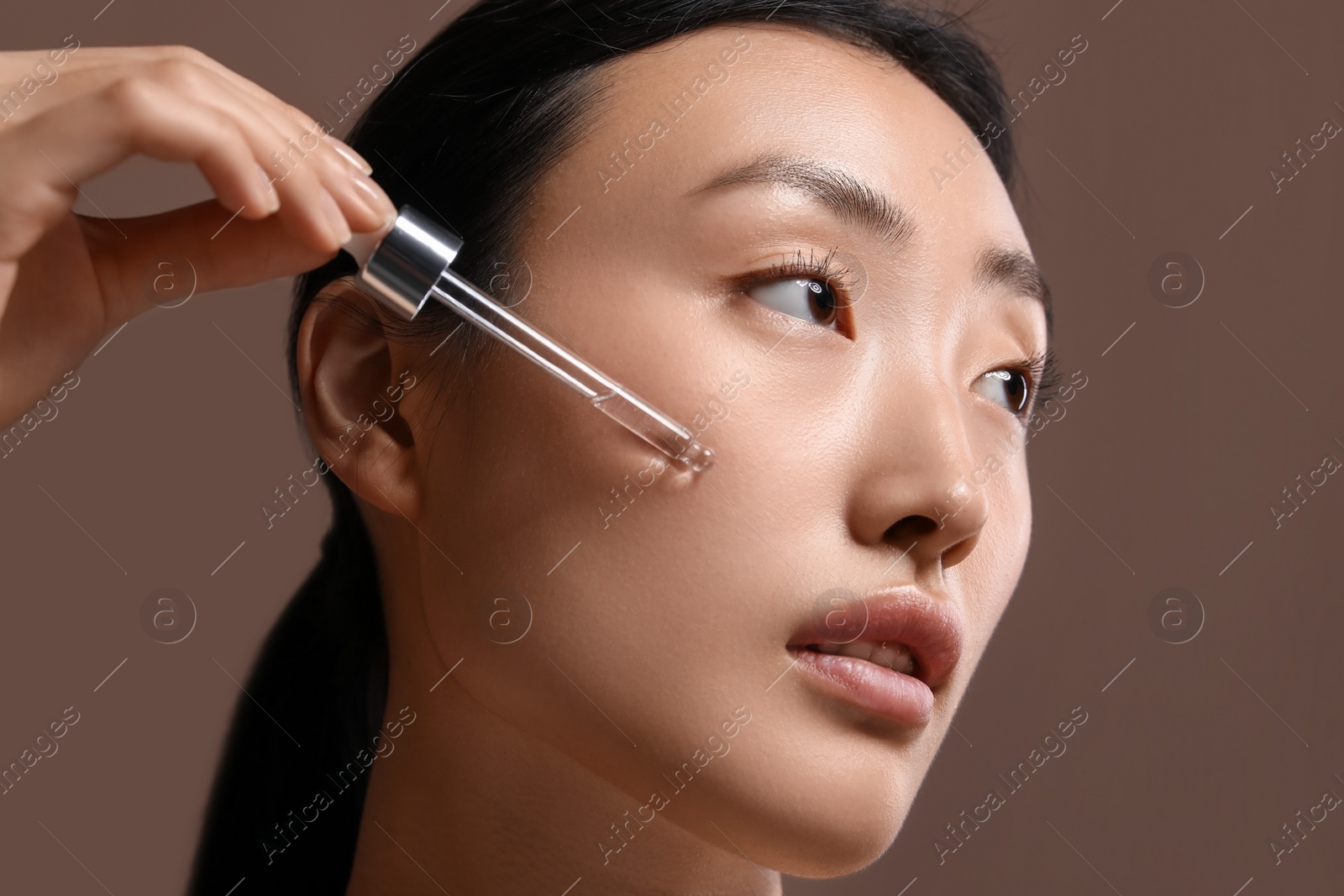 Photo of Beautiful young woman applying cosmetic serum onto her face on brown background, closeup