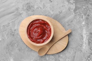 Photo of Organic ketchup in wooden bowl and spoon on grey textured table, top view. Tomato sauce