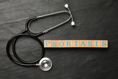 Photo of Word Psoriasis made of wooden cubes with letters and stethoscope on dark gray textured table, flat lay