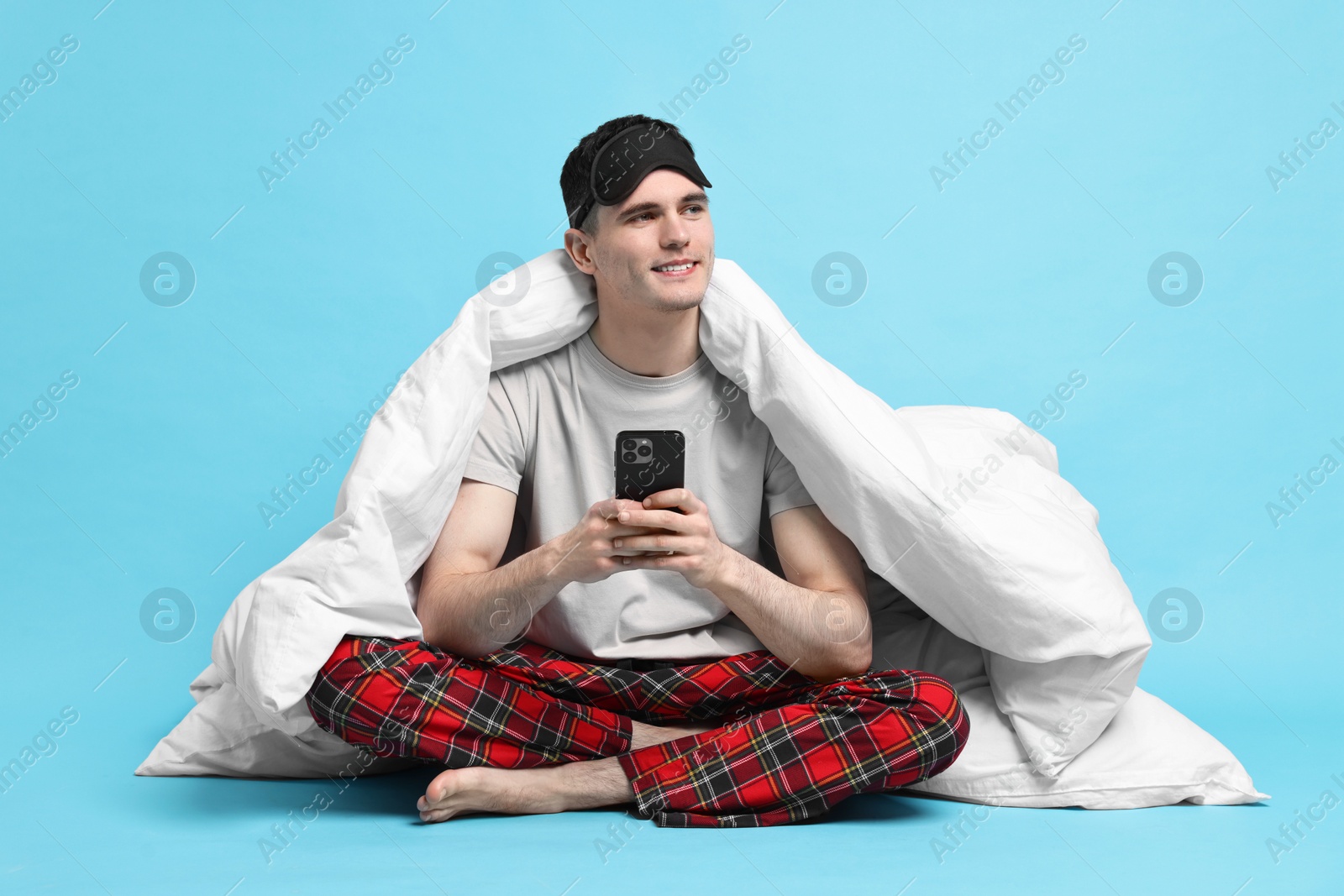 Photo of Happy man in pyjama wrapped in blanket using smartphone on light blue background