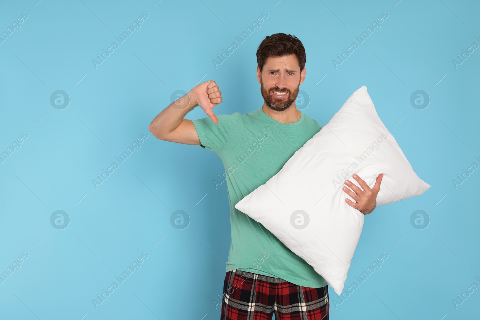 Photo of Sad handsome man showing thumb down and holding soft pillow on light blue background, space for text