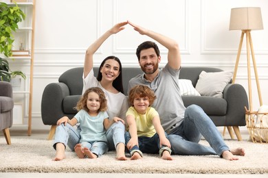Photo of Family housing concept. Happy woman and her husband forming roof with their hands while sitting with kids on floor at home