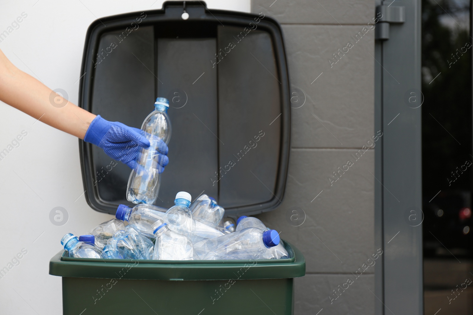 Photo of Woman in gloves putting used plastic bottle into trash bin outdoors, closeup. Recycling problem