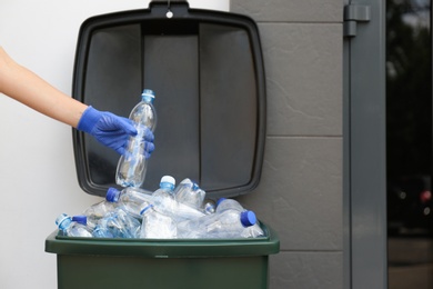 Woman in gloves putting used plastic bottle into trash bin outdoors, closeup. Recycling problem