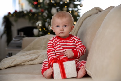 Cute baby in bright Christmas pajamas holding gift box on sofa at home