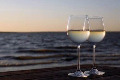 Photo of Two glasses of wine on wooden table near river at sunset. Space for text