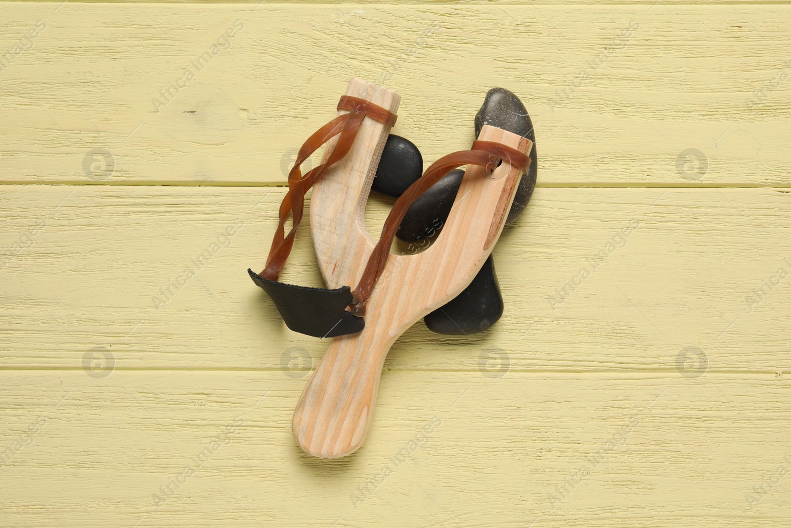 Photo of Slingshot with stones on yellow wooden background, top view