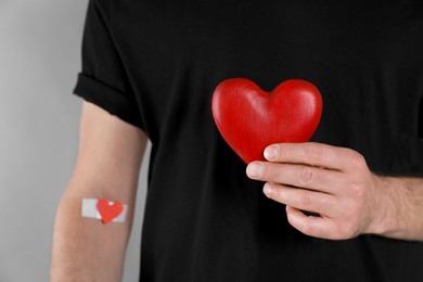 Blood donation concept. Man with adhesive plaster on arm holding red heart against grey background, closeup