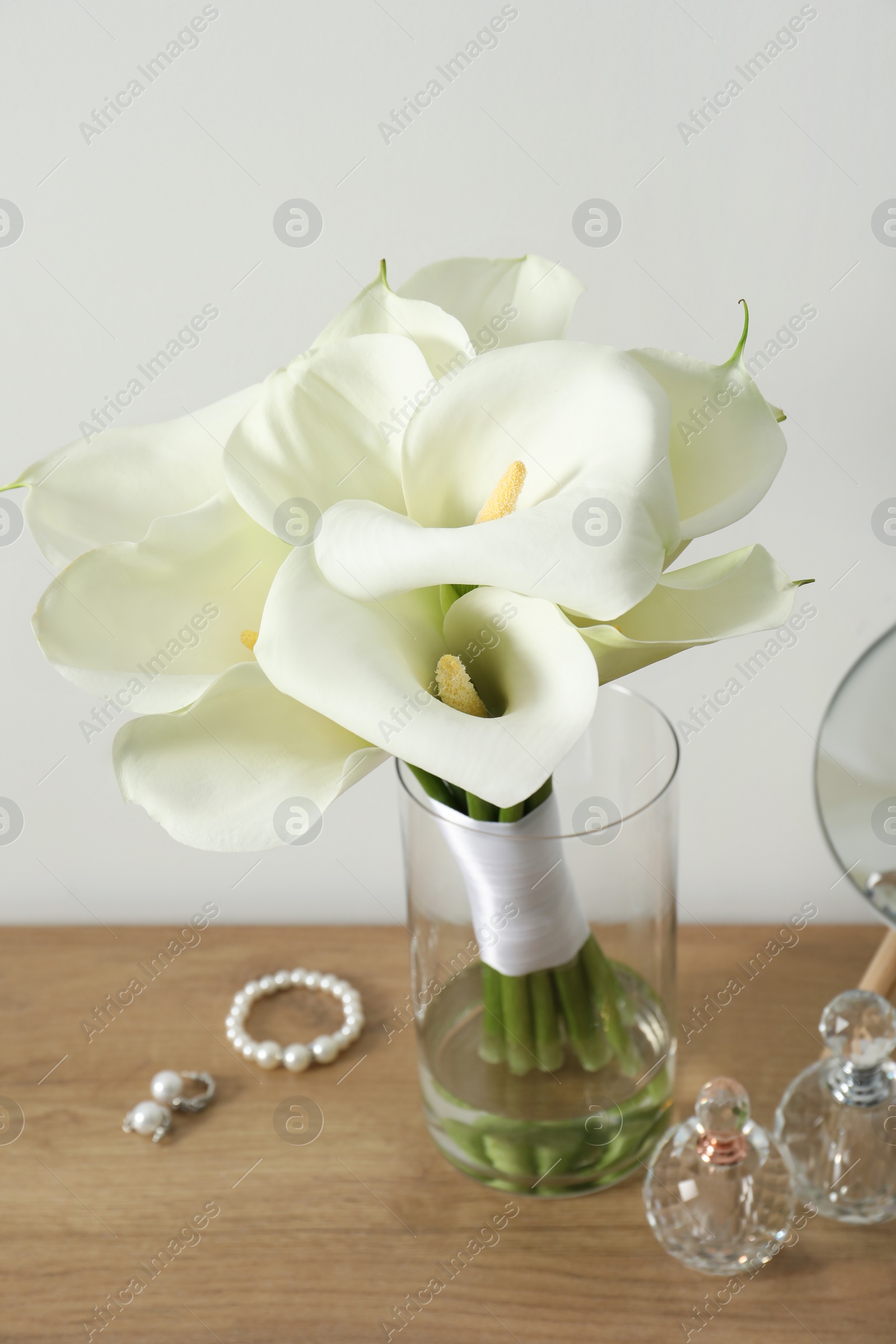 Photo of Beautiful calla lily flowers in glass vase, bottles with perfumes, jewelry and mirror on wooden table