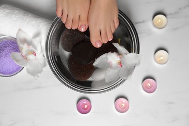 Photo of Woman soaking her feet in bowl with water, flower and spa stones on white marble floor, top view. Pedicure procedure