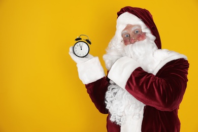 Photo of Santa Claus holding alarm clock on yellow background, space for text. Christmas countdown