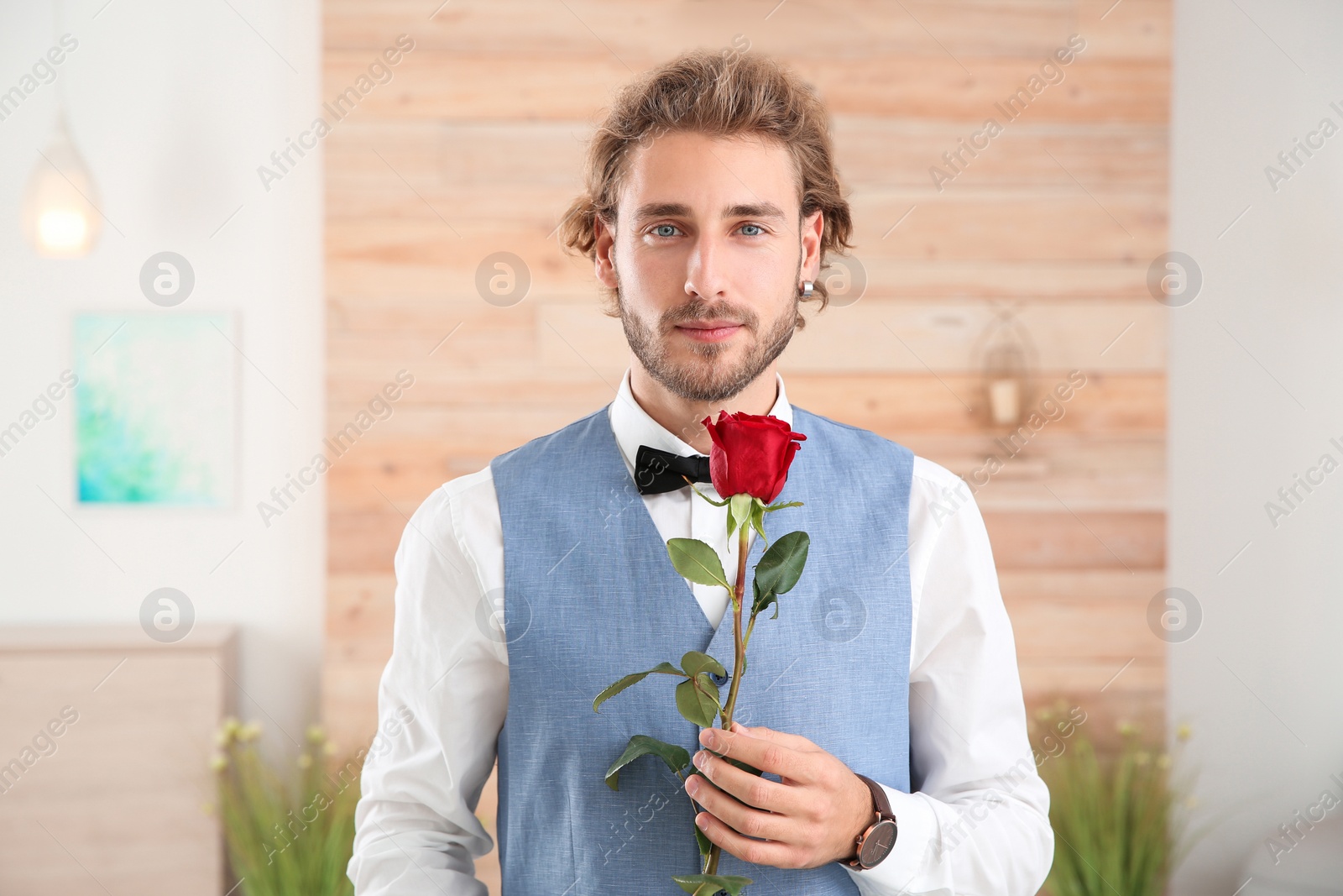 Photo of Handsome man in formal wear holding red rose indoors