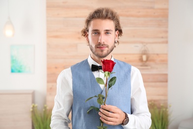 Handsome man in formal wear holding red rose indoors