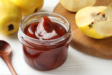 Photo of Tasty homemade quince jam in jar, spoon and fruits on white wooden table, closeup