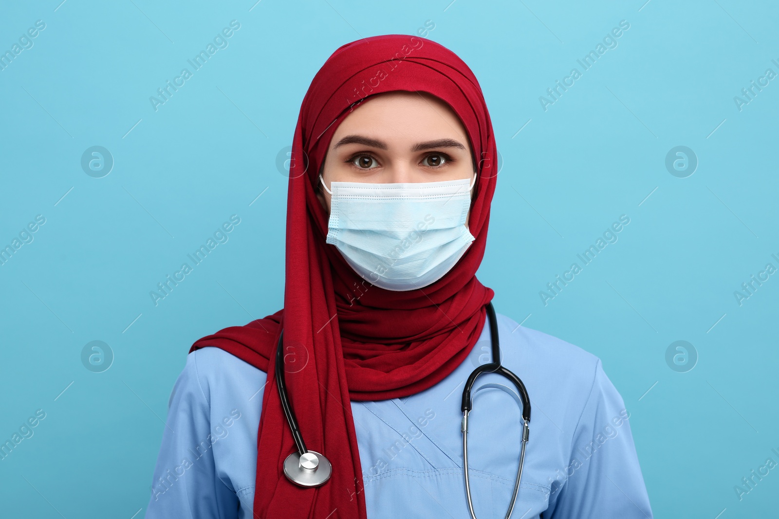 Photo of Muslim woman wearing hijab, medical uniform and protective mask on light blue background