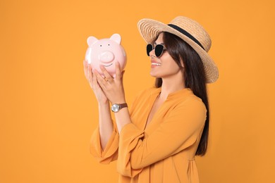 Young woman in stylish sunglasses and straw hat with piggy bank on orange background