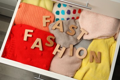 Photo of Phrase FAST FASHION made of wooden letters and different clothes in open drawer, top view