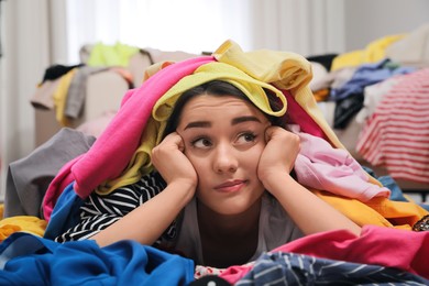 Pensive young woman with lots of clothes on floor in room. Fast fashion