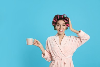 Beautiful young woman in bathrobe with hair curlers holding cup of drink on light blue background