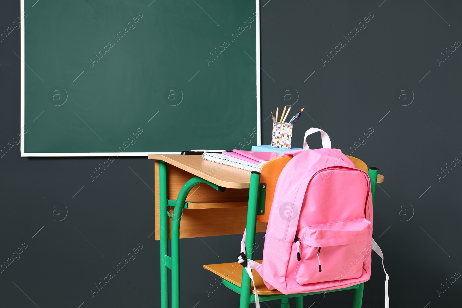 Photo of Wooden school desk with stationery and backpack near blackboard on grey wall