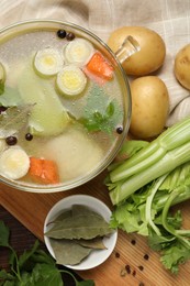 Glass pot with tasty bouillon and different ingredients on wooden table, flat lay