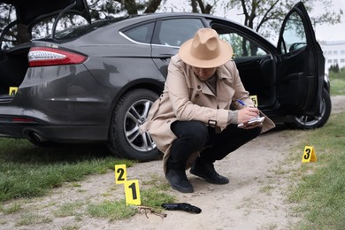Photo of Professional detective in hat examining crime scene near car outdoors