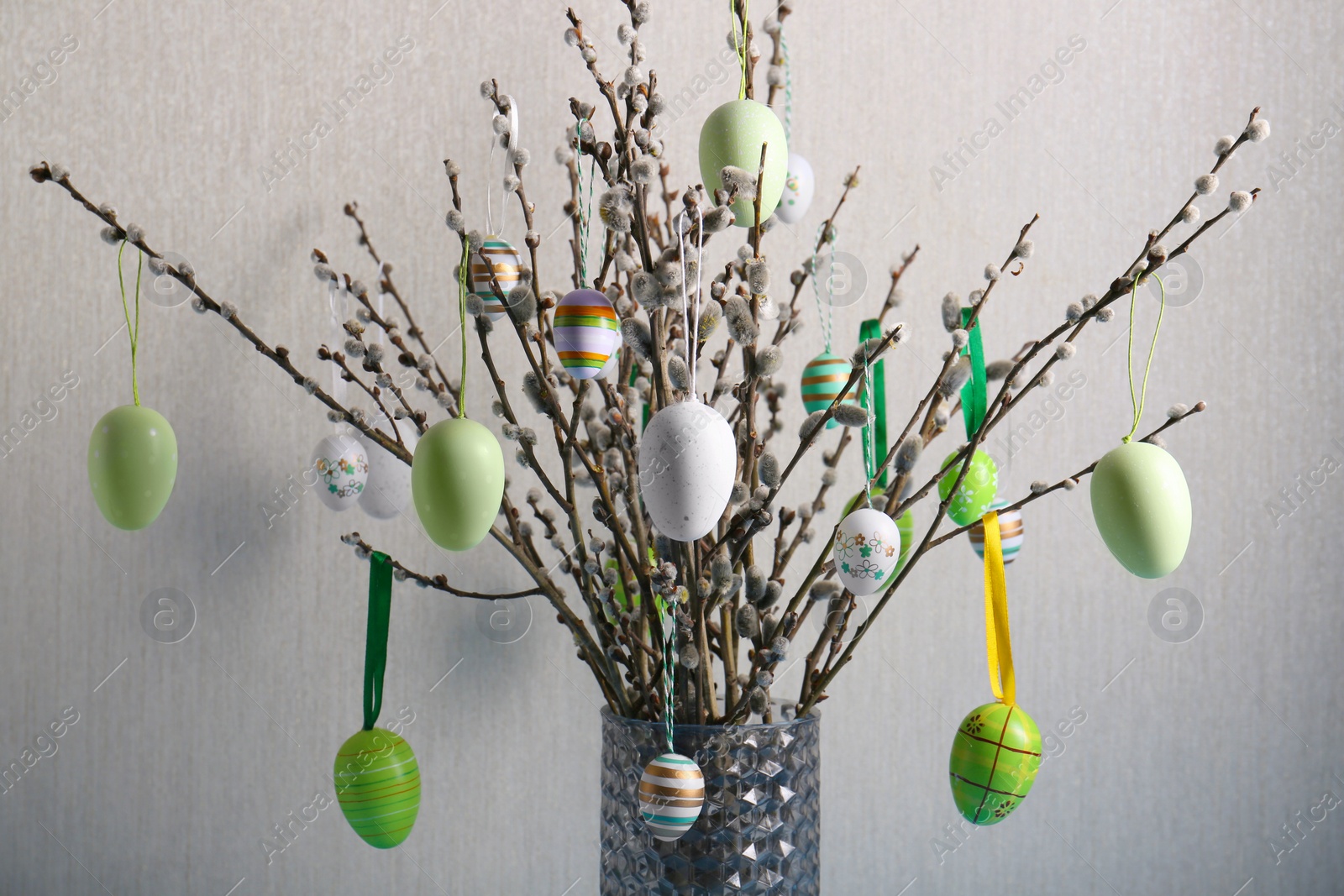 Photo of Beautiful willow branches with painted eggs in vase on light grey background. Easter decor