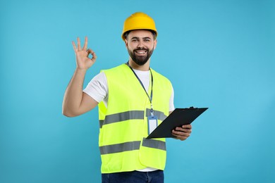 Photo of Engineer in hard hat holding clipboard and showing ok gesture on light blue background