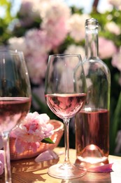 Bottle and glasses of rose wine near beautiful peonies on wooden table in garden