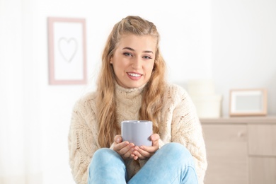 Photo of Attractive young woman in cozy warm sweater with cup of hot drink at home