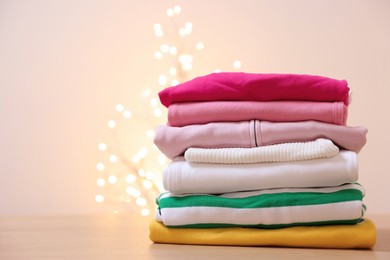 Photo of Stack of folded clothes on wooden table against beige background with blurred lights, space for text