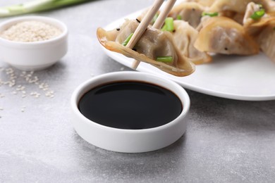 Photo of Holding delicious gyoza (asian dumpling) with chopsticks above soy sauce on light gray table, closeup