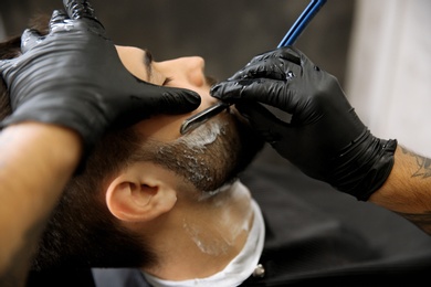 Professional hairdresser shaving client with straight razor in barbershop