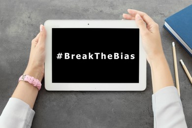 Image of Woman holding tablet with hashtag BreakTheBias on screen at grey table, top view. Campaign theme for International Women's Day
