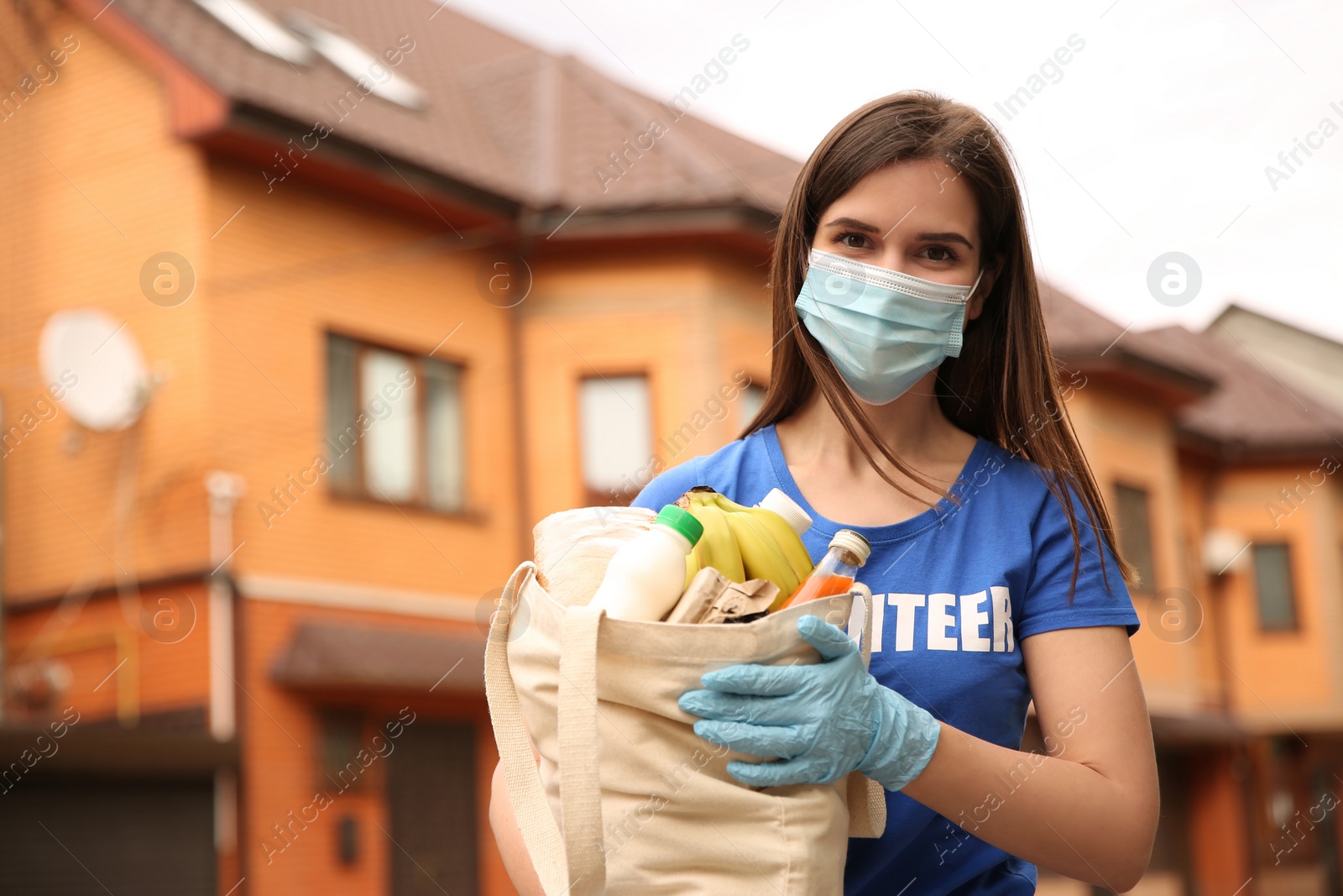 Photo of Female volunteer in protective mask and gloves with products on city street. Aid during coronavirus quarantine