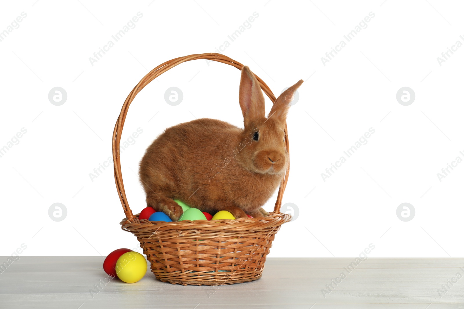 Photo of Cute bunny and basket with Easter eggs on table against white background