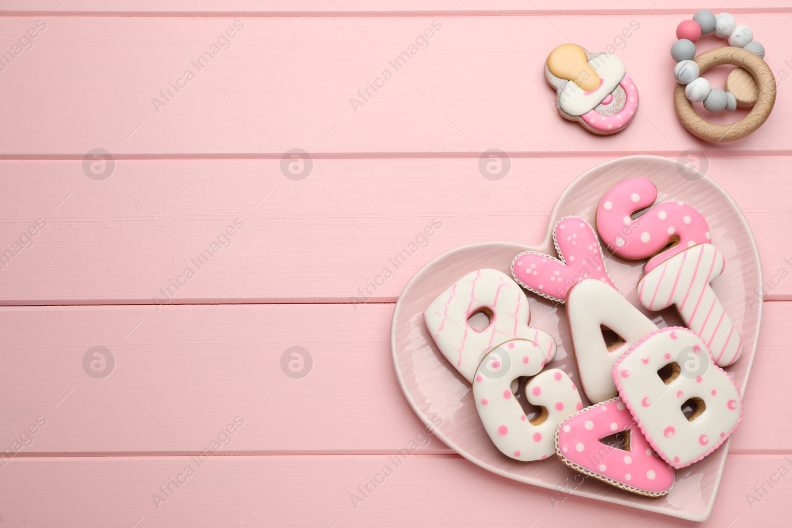 Photo of Heart shaped plate of baby shower cookies and toy on pink wooden table, flat lay. Space for text