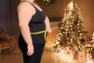 Overweight woman measuring her waist in room with Christmas tree after holidays, closeup