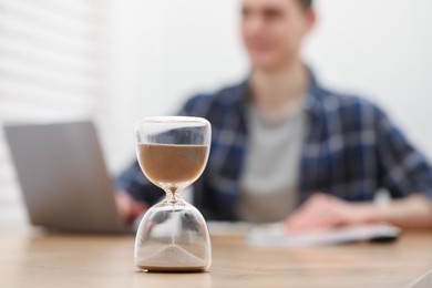 Photo of Hourglass with flowing sand on desk. Man taking notes while using laptop indoors, selective focus