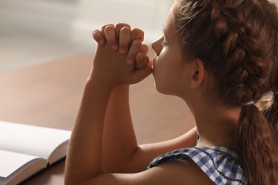 Photo of Cute little girl praying over Bible at table indoors, closeup